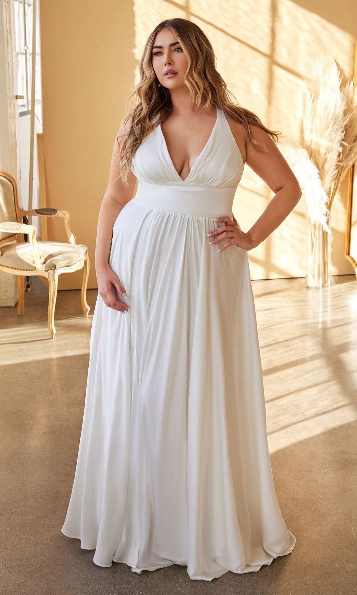 A woman wears the Ladivine 7469WW Long Plus-Size White Dress with a V-neck, standing indoors on a sunlit floor surrounded by neutral furniture and pampas grass.